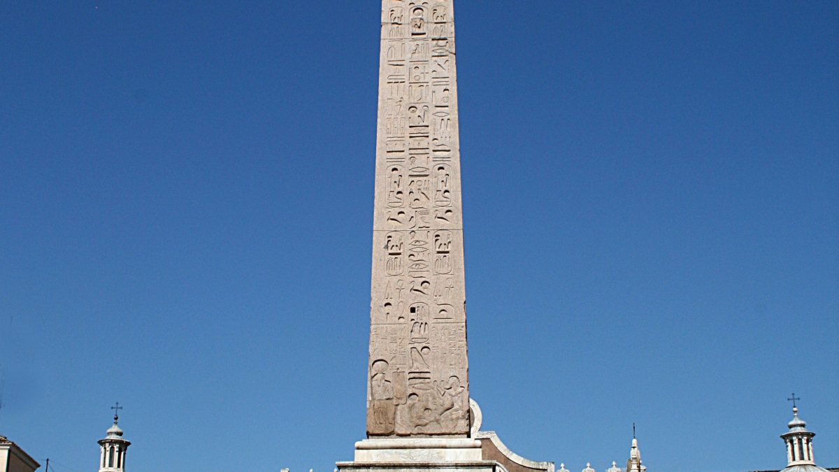 Obelisco Flaminio na Piazza del Popolo em Roma, um monumento antigo egípcio adornado com hieróglifos, sob um céu azul claro. Turistas e locais relaxam e socializam nas escadas na base do obelisco, com a entrada do museu ao fundo.