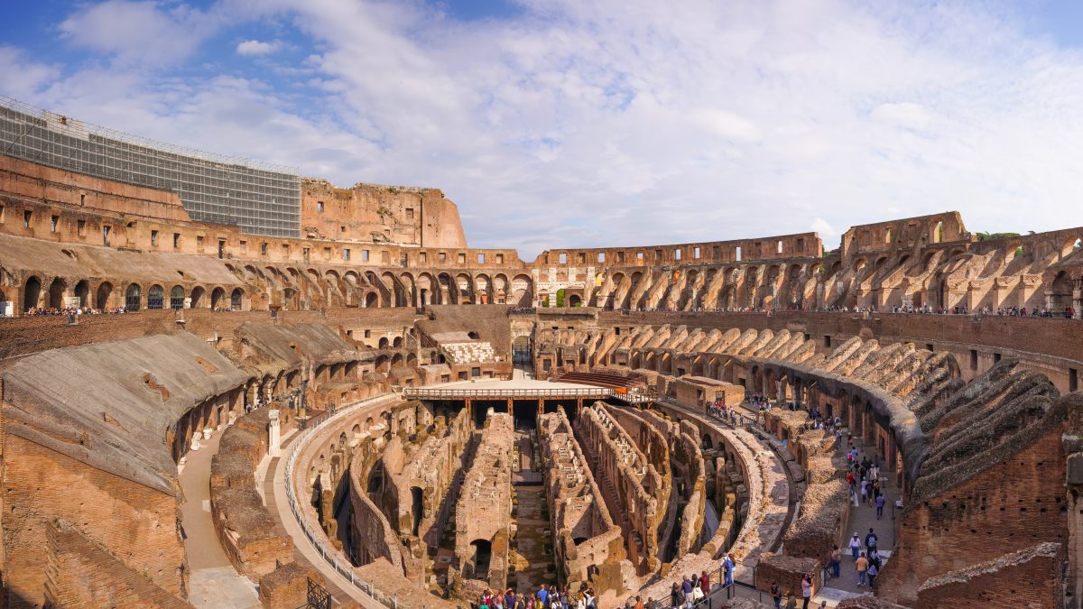 Uma vista panorâmica do interior do Coliseu de Roma, mostrando as ruínas do complexo central e as gradas ao redor, com turistas passeando.