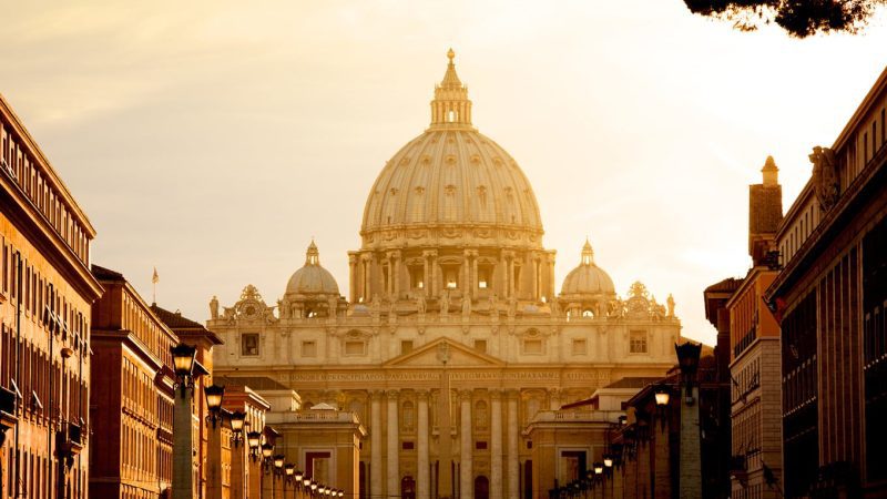 A imagem mostra a Basílica de São Pedro no Vaticano, capturada durante um pôr do sol que banha a construção em uma luz dourada suave.