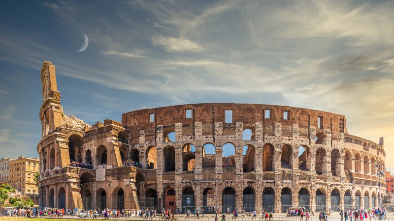 Vista diurna do Coliseu em Roma com turistas ao redor e uma lua crescente visível no céu azul.