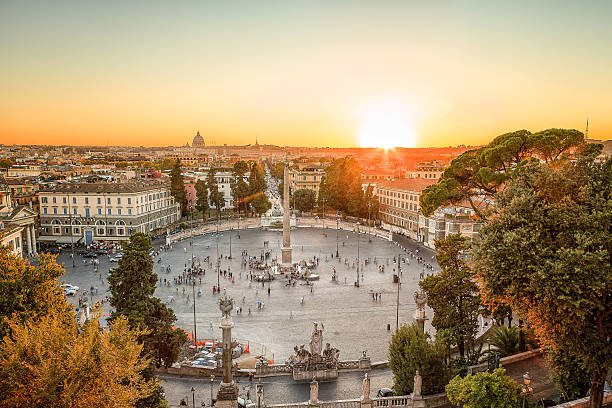 Vista aérea da Piazza del Popolo em Roma ao pôr do sol, destacando o movimentado espaço aberto da praça, o obelisco egípcio Flaminio no centro, fontes e estátuas ornamentadas, e as árvores frondosas. Ao fundo, o céu está tingido de tons quentes de laranja e amarelo enquanto o sol se põe, lançando uma luz dourada sobre a cidade, com vislumbres de monumentos distantes no horizonte.