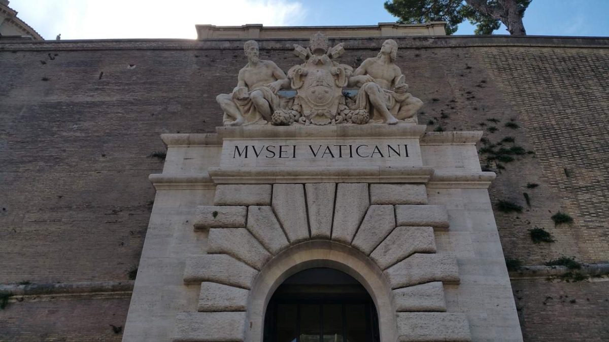 Entrada dos Museus Vaticanos com a inscrição 'MUSEI VATICANI' e esculturas acima do arco."