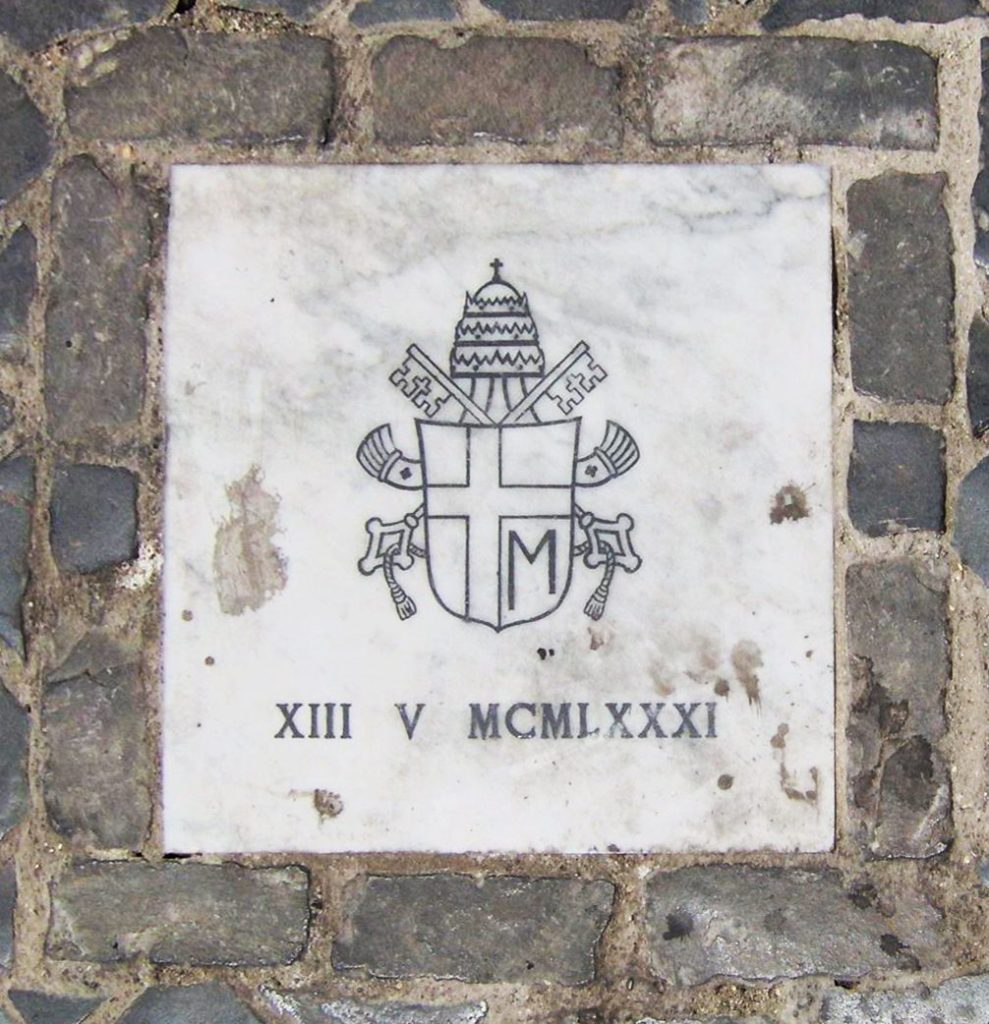 A imagem mostra uma placa comemorativa embutida no chão, cercada por pedras. No centro da placa, há um brasão papal com chaves cruzadas, uma tiara papal e um escudo com a letra "M" na base. Abaixo do brasão, a inscrição "XIII V MCMLXXXI" indica a data 13 de maio de 1981.