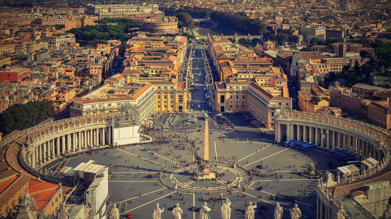 A vista aérea da Praça de São Pedro em Roma revela uma impressionante exibição de simetria arquitetônica: um colossal obelisco egípcio ergue-se ao centro, flanqueado por colunatas curvas e a silhueta histórica da cidade ao fundo.