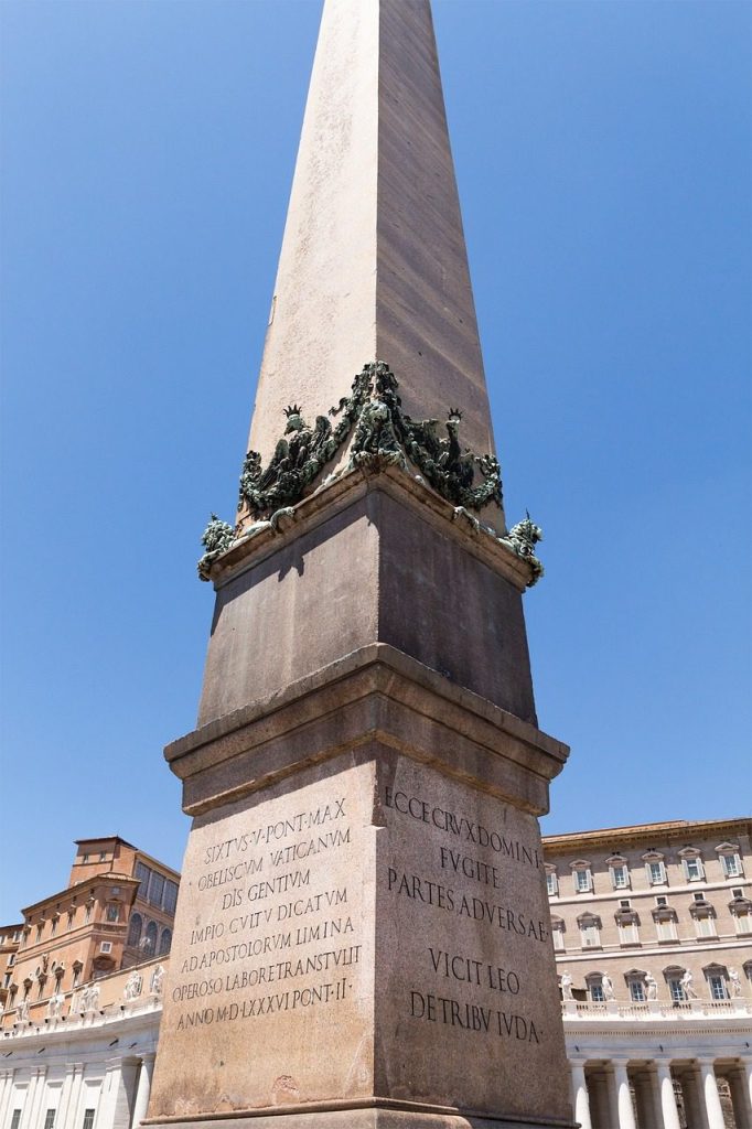 Um obelisco alto se ergue contra um céu azul límpido, com inscrições em latim em sua base, situado na Praça de São Pedro no Vaticano, ladeado pela arquitetura simétrica das colunatas ao fundo.
