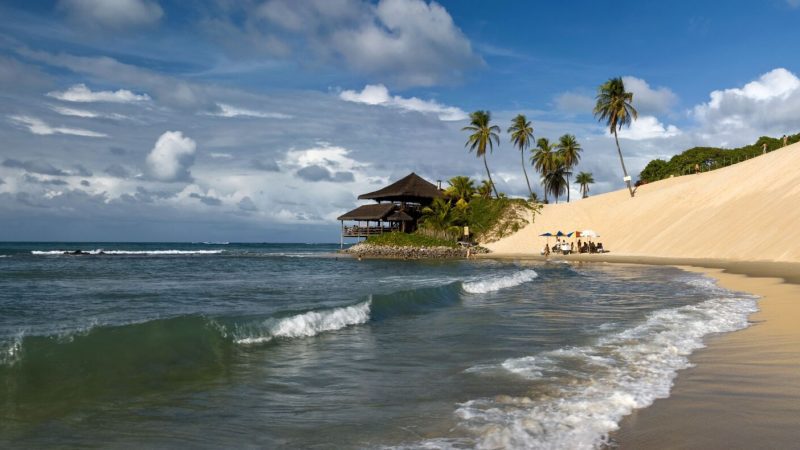 Praia de Genipabu, em Natal, Rio Grande do Norte, com suas icônicas dunas de areia branca, coqueiros e uma estrutura à beira-mar. As ondas calmas e o céu azul complementam a paisagem paradisíaca.