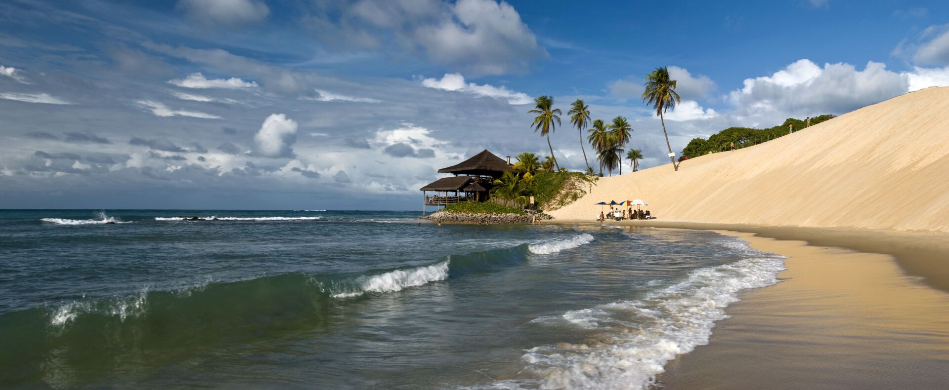 Praia de Genipabu, em Natal, Rio Grande do Norte, com suas icônicas dunas de areia branca, coqueiros e uma estrutura à beira-mar. As ondas calmas e o céu azul complementam a paisagem paradisíaca.