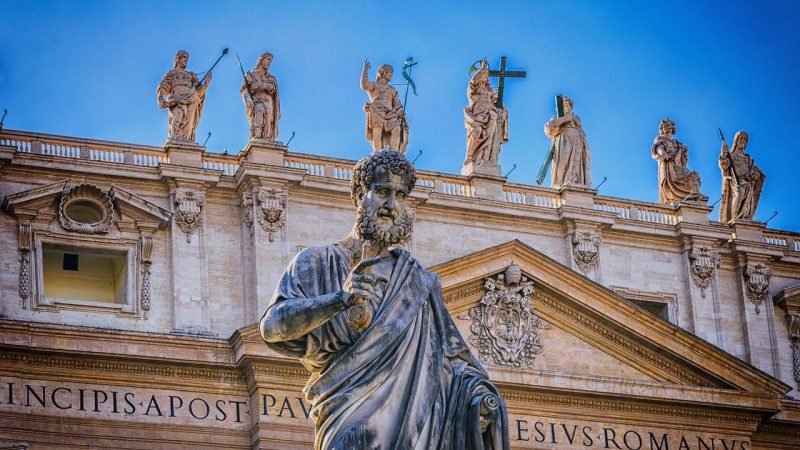 A imagem mostra uma estátua de São Pedro com uma chave na mão, em primeiro plano, diante da Basílica de São Pedro no Vaticano. Ele é apresentado com uma expressão séria e um dedo apontando para cima, destacando-se contra o céu azul claro.