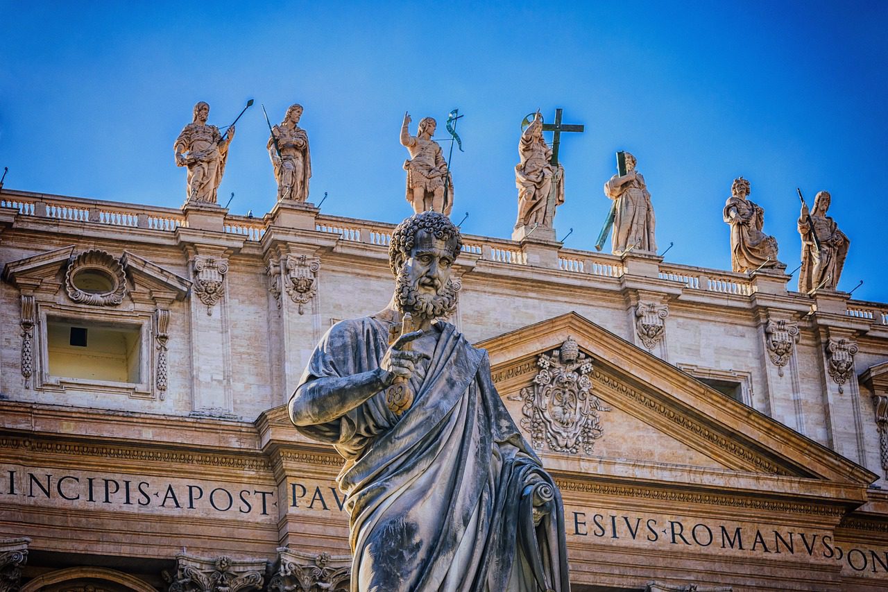 A imagem mostra uma estátua de São Pedro com uma chave na mão, em primeiro plano, diante da Basílica de São Pedro no Vaticano. Ele é apresentado com uma expressão séria e um dedo apontando para cima, destacando-se contra o céu azul claro.