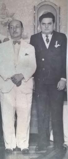 Foto antiga de dois homens posando juntos, ambos vestindo ternos elegantes; um de branco e outro de preto.