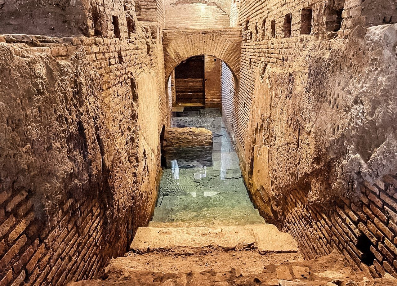 A imagem mostra uma estrutura subterrânea de tijolos antigos, caracterizada por paredes robustas e um teto arqueado. Há uma escada de pedra que leva a uma área mais baixa, parcialmente submersa em água cristalina. A arquitetura sugere uma construção histórica, possivelmente romana, com detalhes de alvenaria bem preservados. A luz natural ilumina o espaço, destacando as texturas das paredes e a transparência da água. Este local é provavelmente uma parte do subterrâneo da Fontana di Trevi