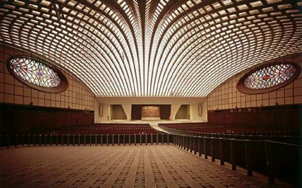 Interior da Sala Paulo VI no Vaticano, com seu impressionante teto em forma de ondas e grandes vitrais ovais nas laterais. O espaço é usado para audiências papais e eventos especiais.