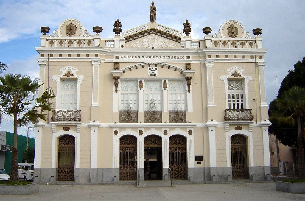 Fachada do Teatro Alberto Maranhão, um edifício histórico com arquitetura eclética, localizado em Natal, Rio Grande do Norte. A construção exibe detalhes ornamentais elaborados e varandas decorativas, com palmeiras ao redor.