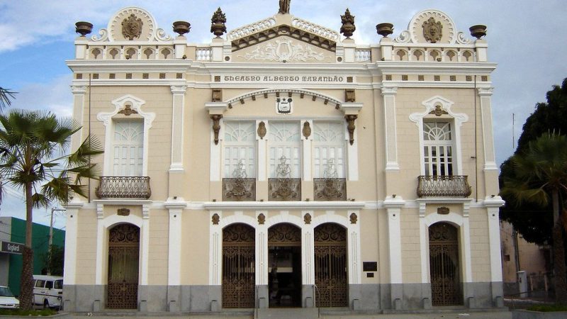 Fachada do Teatro Alberto Maranhão, um edifício histórico com arquitetura eclética, localizado em Natal, Rio Grande do Norte. A construção exibe detalhes ornamentais elaborados e varandas decorativas, com palmeiras ao redor.