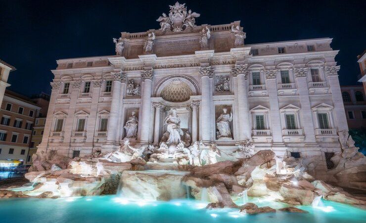 A imagem exibe a majestosa Fontana di Trevi, um dos monumentos mais icônicos de Roma, Itália. À noite, a fonte brilha com iluminação que destaca suas impressionantes esculturas barrocas e a beleza arquitetônica detalhada, criando uma atmosfera mágica e encantadora.