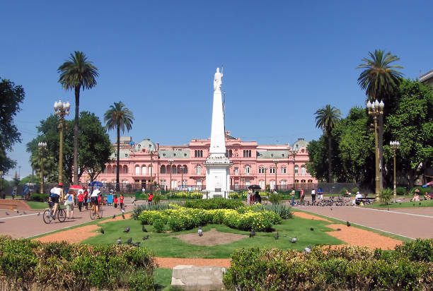 A imagem mostra a Casa Rosada, sede do governo argentino, localizada na Plaza de Mayo, em Buenos Aires. Em frente à Casa Rosada, há um obelisco branco cercado por jardins bem cuidados, com palmeiras e pessoas passeando pela praça.