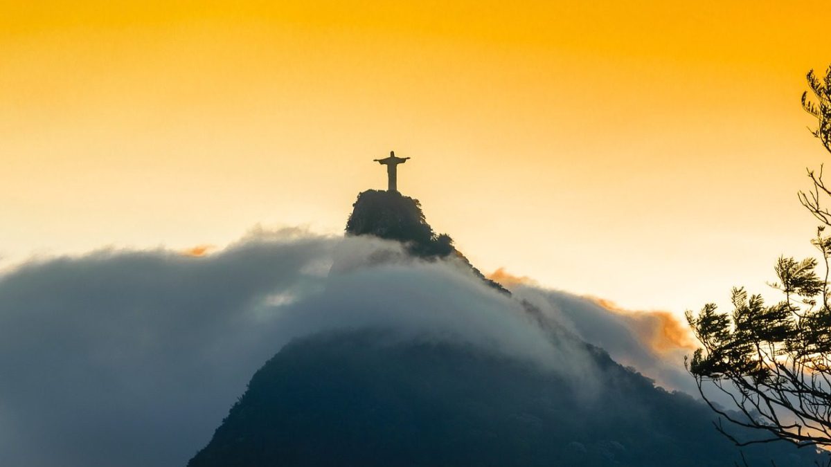 A estátua está em silhueta contra um céu alaranjado, sugerindo nascer ou pôr do sol. As nuvens envolvem a base da montanha, dando à cena uma aparência mística ou etérea. A estátua é icónica e fica no topo do monte Corcovado, com vista para a cidade. A imagem captura um momento tranquilo e poderoso, destacando a grandiosidade da estátua e a beleza natural do seu entorno. Não há texto na imagem, apenas a paisagem natural e a estátua.