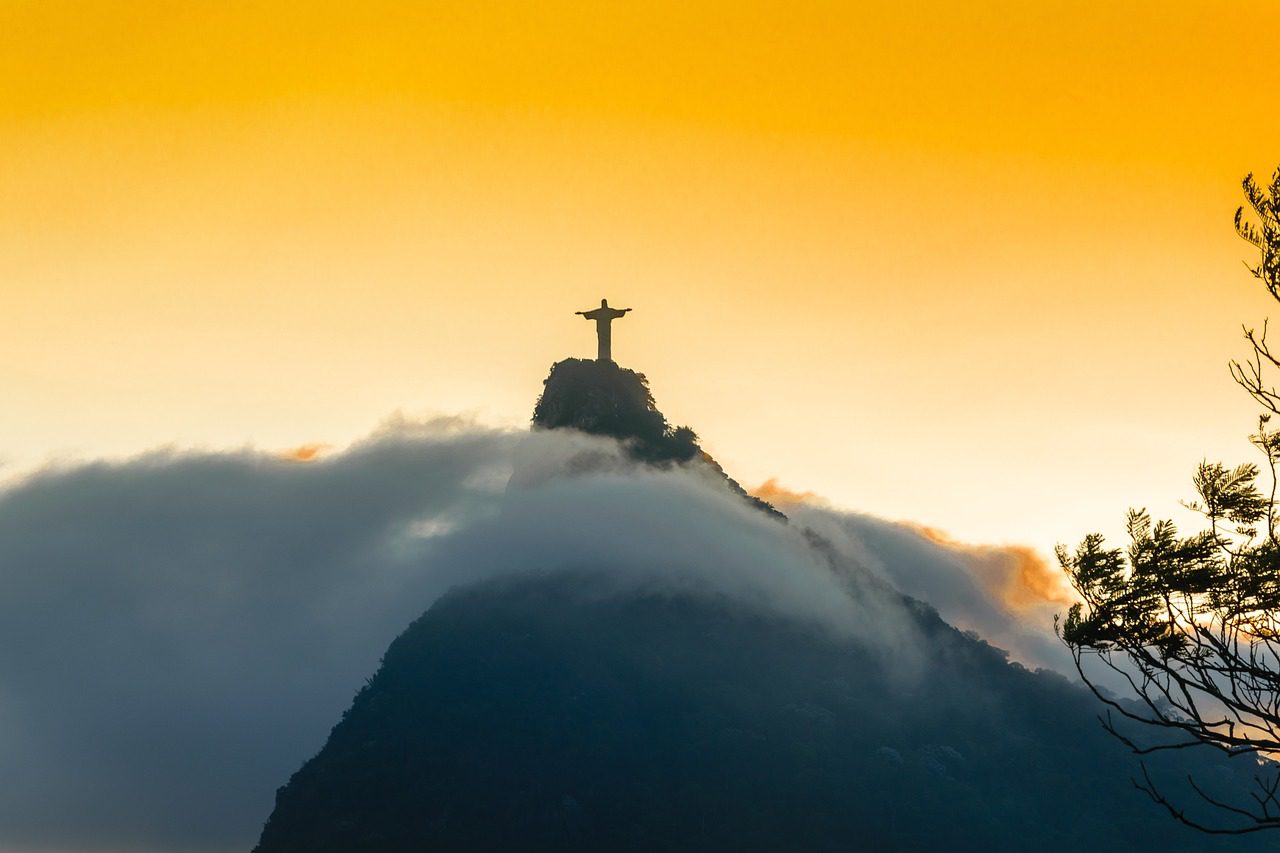 A estátua está em silhueta contra um céu alaranjado, sugerindo nascer ou pôr do sol. As nuvens envolvem a base da montanha, dando à cena uma aparência mística ou etérea. A estátua é icónica e fica no topo do monte Corcovado, com vista para a cidade. A imagem captura um momento tranquilo e poderoso, destacando a grandiosidade da estátua e a beleza natural do seu entorno. Não há texto na imagem, apenas a paisagem natural e a estátua.