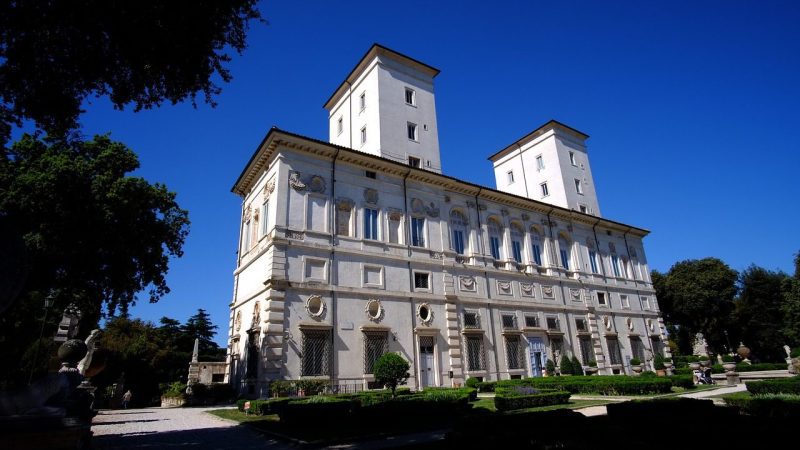 Imagem da Galeria Borghese em Roma, destacando sua arquitetura renascentista imponente com duas torres e detalhes ornamentais, rodeada por jardins bem cuidados em um dia de céu azul claro.