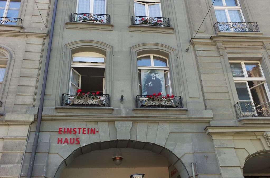 A fachada da Casa de Einstein em Berna, com a inscrição "Einstein-Haus" em vermelho, apresenta janelas decoradas com flores vermelhas e uma imagem de Einstein visível na janela central. Esta casa histórica exibe a vida e o trabalho de Albert Einstein durante seu período em Berna.