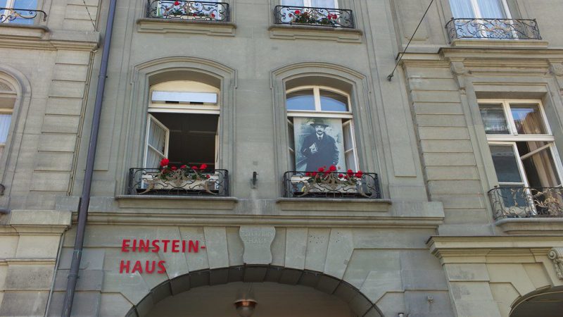 A fachada da Casa de Einstein em Berna, com a inscrição "Einstein-Haus" em vermelho, apresenta janelas decoradas com flores vermelhas e uma imagem de Einstein visível na janela central. Esta casa histórica exibe a vida e o trabalho de Albert Einstein durante seu período em Berna.