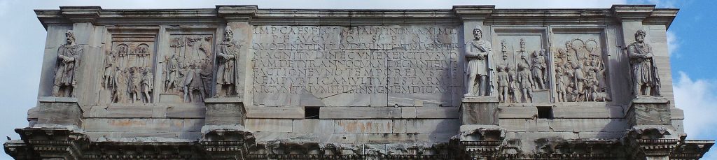 Close-up da Ática do Arco de Constantino em Roma. Esta seção apresenta várias esculturas e inscrições. O painel central possui uma inscrição em latim comemorando a vitória de Constantino e sua dedicação ao povo romano. Flanqueando esta inscrição central, há vários relevos esculpidos que retratam diferentes cenas e estátuas de soldados romanos ou imperadores.