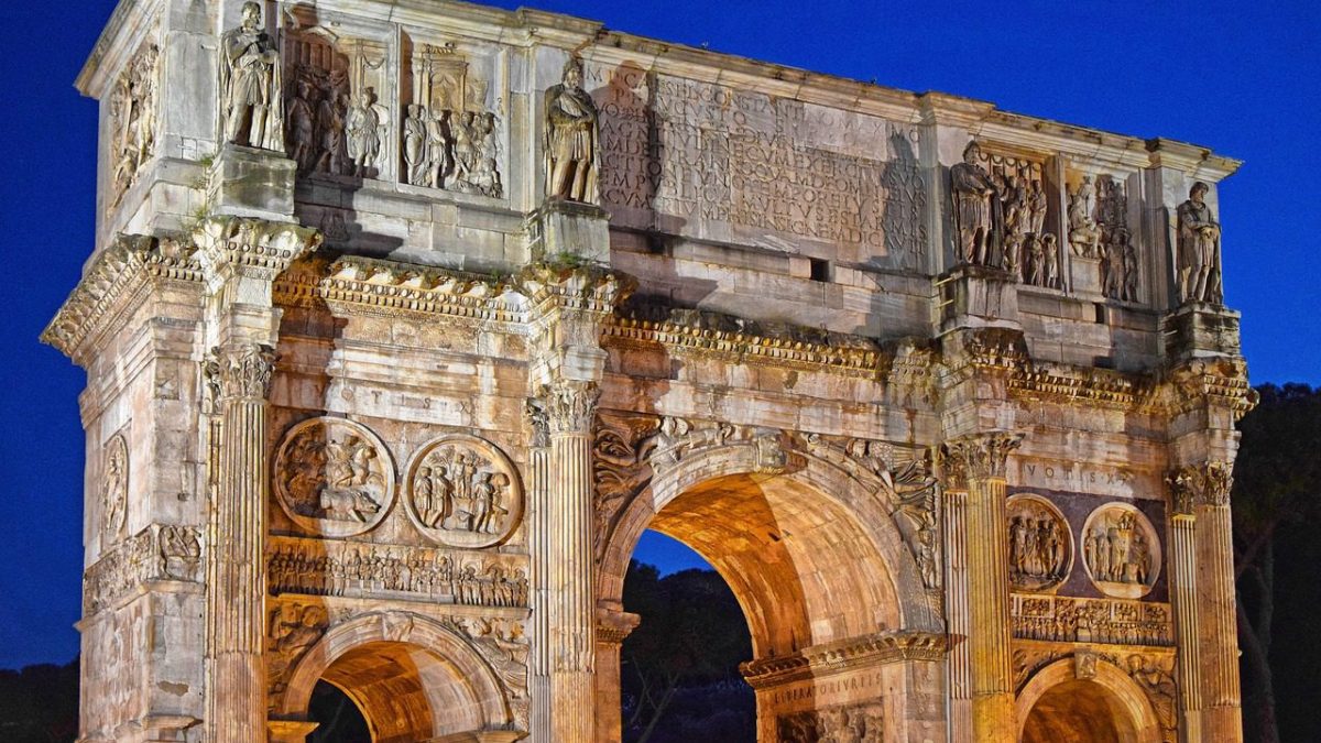 O Arco de Constantino em Roma, iluminado à noite, destaca suas esculturas e relevos detalhados. No topo, uma inscrição central é ladeada por relevos de Marco Aurélio. Abaixo, medalhões da época de Adriano e Constantino e frisos com cenas das vitórias de Constantino.