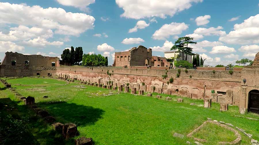 Vista panorâmica das ruínas do Monte Palatino em Roma, Itália, com arcos antigos, vegetação e estruturas históricas ao fundo.