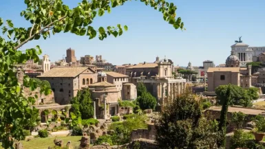 Essa imagem mostra uma vista panorâmica das ruínas antigas de Roma, incluindo diversos templos, arcos e estruturas históricas, cercados por vegetação e com a cidade moderna ao fundo. As ruínas são parte do Fórum Romano, um dos locais mais emblemáticos da história romana.