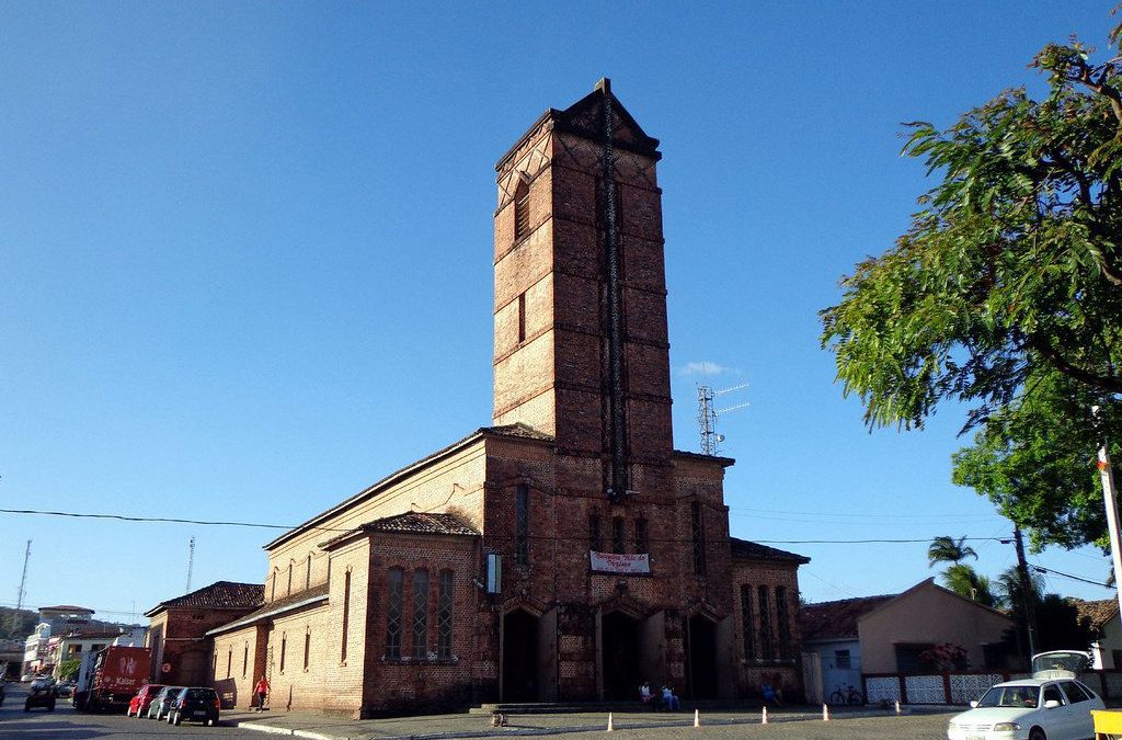 A imagem mostra a Igreja Santa Rita de Cássia em Rio Tinto, uma construção em tijolos aparentes com uma imponente torre central. A arquitetura reflete influências alemãs, com linhas simples e uma aparência robusta. A praça em frente à igreja, onde se encontra a estátua do fundador da cidade, Coronel Frederico João Lundgren, também é visível.