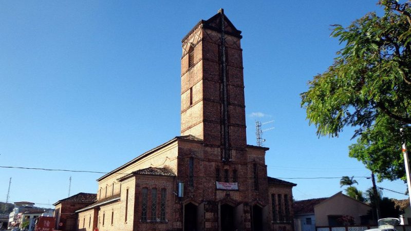 A imagem mostra a Igreja Santa Rita de Cássia em Rio Tinto, uma construção em tijolos aparentes com uma imponente torre central. A arquitetura reflete influências alemãs, com linhas simples e uma aparência robusta. A praça em frente à igreja, onde se encontra a estátua do fundador da cidade, Coronel Frederico João Lundgren, também é visível.