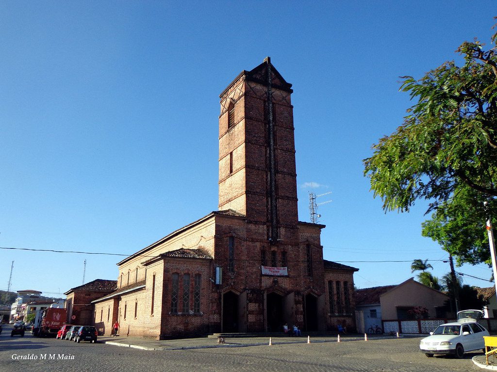 A imagem mostra a Igreja Santa Rita de Cássia em Rio Tinto, uma construção em tijolos aparentes com uma imponente torre central. A arquitetura reflete influências alemãs, com linhas simples e uma aparência robusta. A praça em frente à igreja, onde se encontra a estátua do fundador da cidade, Coronel Frederico João Lundgren, também é visível.