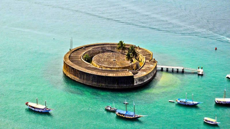 Imagem aérea do Forte de São Marcelo, localizado na Baía de Todos os Santos, em Salvador. O forte tem formato circular e está rodeado por águas esverdeadas. Algumas embarcações estão ancoradas próximas à fortificação.