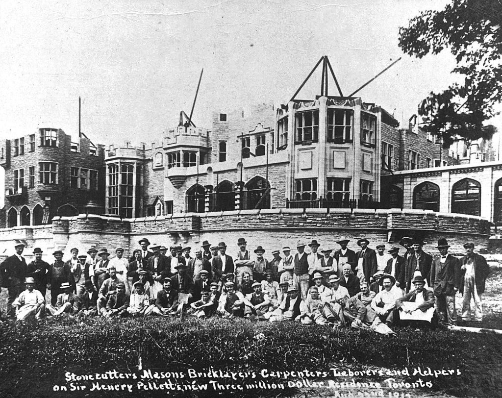 Foto histórica da construção da Casa Loma, em Toronto, por volta de 1912, com um grupo de pedreiros, carpinteiros e trabalhadores posando em frente ao edifício em construção. A imagem mostra o progresso das obras da icônica mansão de Sir Henry Pellatt. O castelo, ainda incompleto, já exibe suas imponentes torres e fachada em estilo gótico.