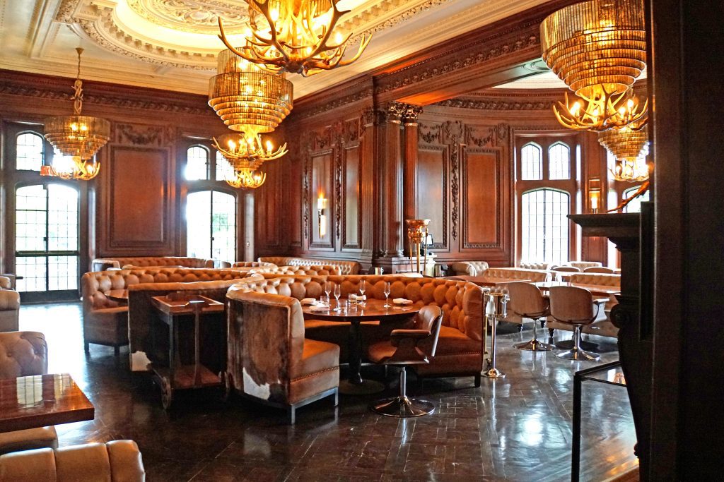 Interior luxuoso da Casa Loma, com decoração clássica em madeira escura e lustres sofisticados. O ambiente elegante conta com sofás de couro e mesas redondas, criando uma atmosfera refinada e acolhedora. A luz suave realça os detalhes esculpidos das paredes e teto, proporcionando uma experiência única.