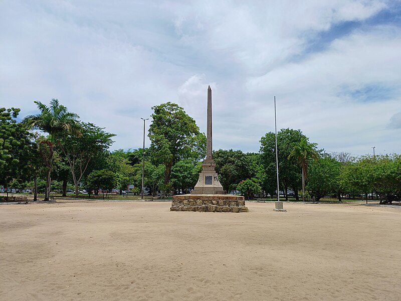 Obelisco central em uma praça arborizada, cercado por árvores e um amplo espaço de areia. O céu está parcialmente nublado, destacando a estrutura do obelisco, que é um ponto focal no ambiente. Ao redor, há uma vegetação abundante que contrasta com o solo claro.