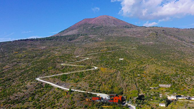 A imagem mostra o Monte Vesúvio ao fundo, com uma estrada sinuosa que sobe a montanha, cortando a vegetação esparsa ao longo da encosta. Algumas casas, incluindo uma destacada em vermelho, estão localizadas próximas à base do vulcão. O céu azul e as nuvens leves complementam a paisagem, destacando a imponência do Vesúvio e o percurso que leva até sua cratera.