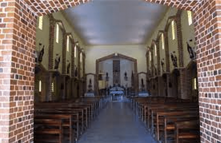 Interior da Igreja de Santa Rita em Rio Tinto, com bancos de madeira dispostos em fileiras que levam ao altar ao fundo. As paredes laterais são adornadas com estátuas de santos e possuem grandes janelas estreitas que iluminam o espaço com luz natural. A arquitetura interna reflete o estilo simples e acolhedor da igreja.