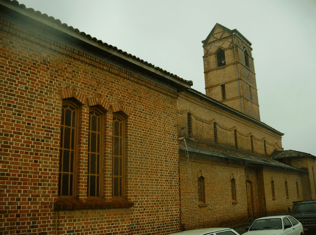 A imagem mostra uma vista lateral da Igreja Santa Rita de Cássia em Rio Tinto, destacando a construção em tijolos aparentes e as janelas emolduradas por arcos de tijolos. A torre central se eleva imponente, complementando a estrutura sólida e austera, característica da arquitetura com influência alemã.