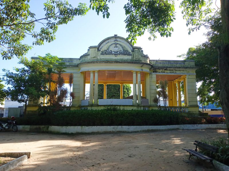 Coreto antigo de arquitetura clássica, com colunas e detalhes ornamentados, localizado em uma praça arborizada. A estrutura amarela está rodeada por vegetação e um caminho de terra, proporcionando um ambiente sereno. A luz do dia ilumina a fachada, destacando os detalhes da construção.