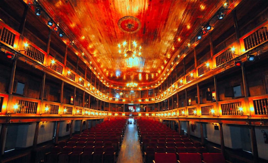 A imagem mostra o interior do Theatro Santa Roza, localizado em João Pessoa, Paraíba, um dos teatros mais antigos do Brasil. Com arquitetura italiana e grega o espaço destaca-se pelos detalhes em madeira, lustres imponentes e varandas que circundam o salão. O teatro é um importante patrimônio cultural e histórico da cidade, oferecendo um ambiente acolhedor para apresentações artísticas.