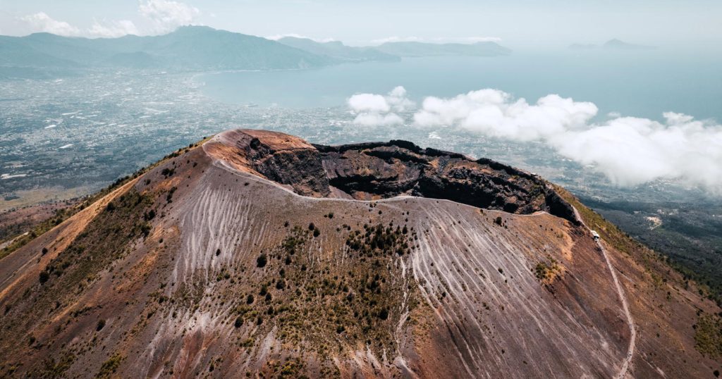 A imagem mostra a cratera do Monte Vesúvio, com sua forma impressionante e suas encostas cobertas de vegetação e marcas vulcânicas. Ao fundo, é possível ver o Golfo de Nápoles e a cidade, parcialmente cobertos por nuvens, destacando o contraste entre a natureza e a vida urbana. O cenário transmite a grandiosidade e a força natural do vulcão, que é um dos mais icônicos do mundo.