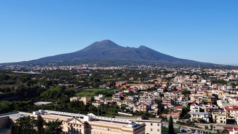 A imagem mostra uma vista panorâmica do Monte Vesúvio, com seu imponente cume vulcânico ao fundo, cercado por uma vasta área urbana. A cidade se estende pela planície, com casas, edifícios e vegetação espalhados, enquanto o céu está limpo, destacando o contraste entre a modernidade da cidade e a natureza poderosa do vulcão.