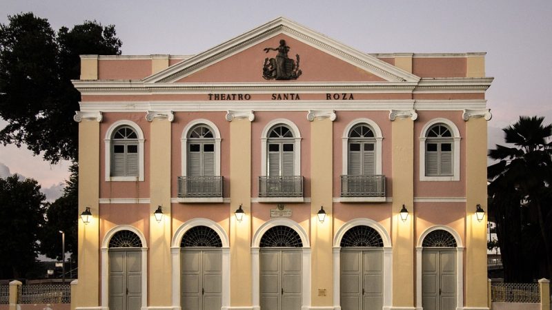 A imagem mostra a fachada do Theatro Santa Roza, em João Pessoa, com sua arquitetura italiana preservada. A estrutura destaca elementos como colunas simétricas e arcos, além do frontão triangular no topo, que mantém o charme histórico do teatro, inaugurado em 1889.