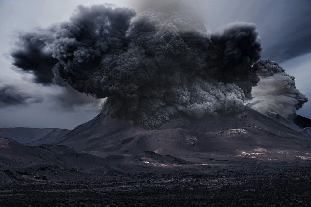 A imagem mostra um vulcão em erupção, com uma imensa nuvem de fumaça escura e cinzas sendo expelida de sua cratera. A paisagem ao redor é árida e coberta por rochas vulcânicas, enquanto a fumaça densa se espalha pelo céu, criando uma atmosfera dramática e poderosa. A cena transmite a força e o impacto devastador de uma erupção vulcânica.